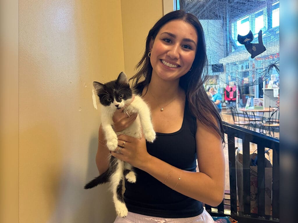 A young woman with long brown hair smiles while holding a small black and white kitten in her arms. The background features a bright room with various items, likely an adoption center or pet shop.