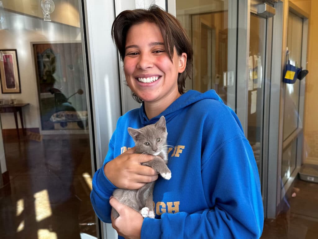 A young woman smiling while holding a small gray kitten in her arms. She is wearing a blue sweatshirt and standing in a brightly lit indoor space with glass doors in the background. The setting appears welcoming and casual.