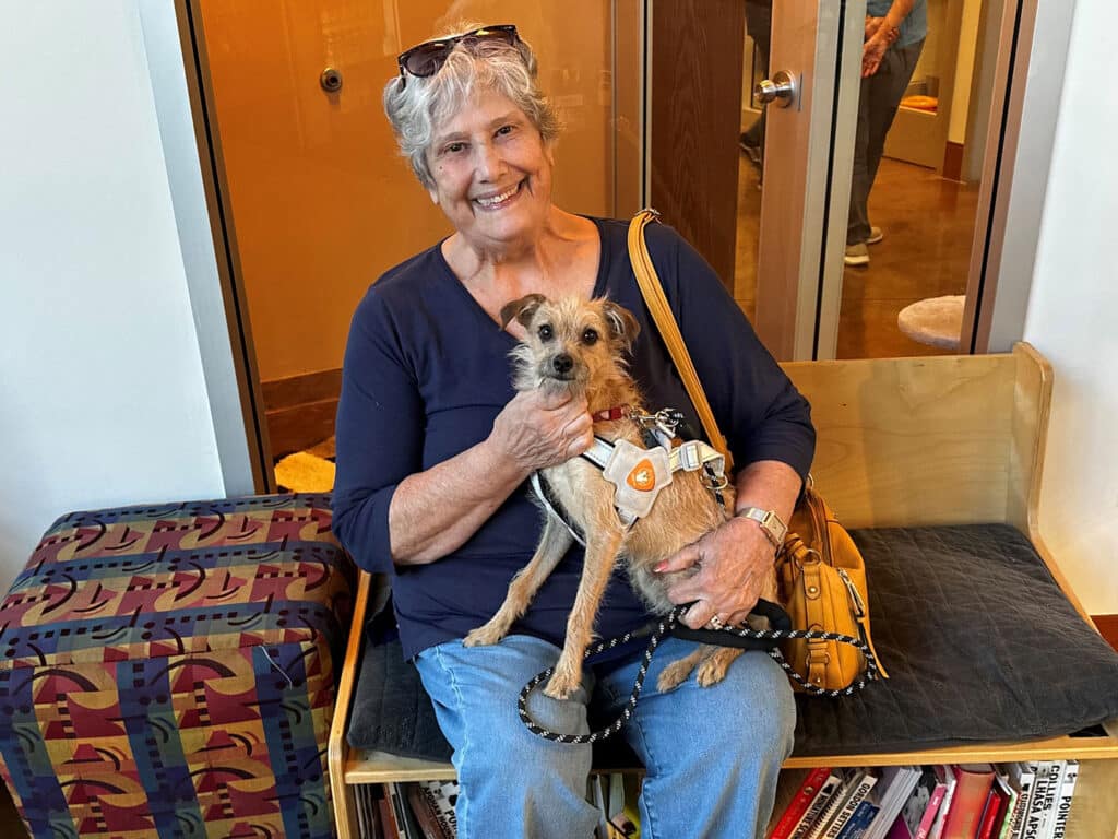 A smiling older woman with gray hair sits on a bench holding a small dog wearing a harness. The background features a wooden wall and an open doorway, indicating an indoor space. The woman wears a navy top and light blue jeans.