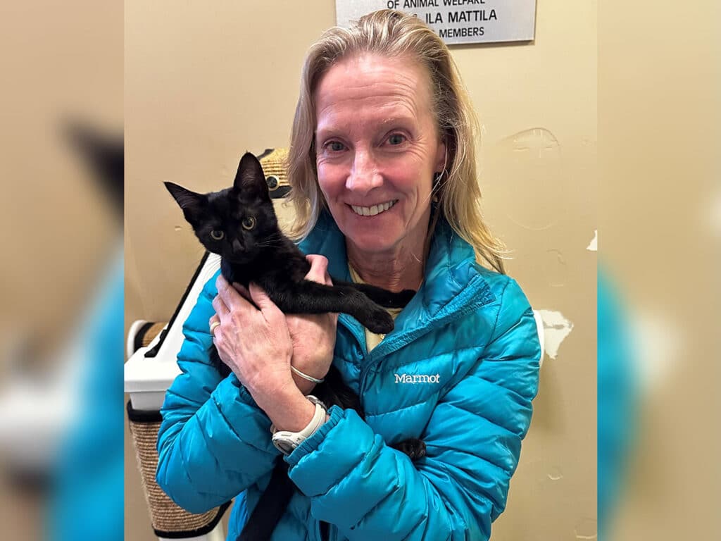 A woman wearing a turquoise jacket holds a small black kitten in her arms, smiling. The background features a sign related to animal welfare, creating a setting that highlights pet adoption.
