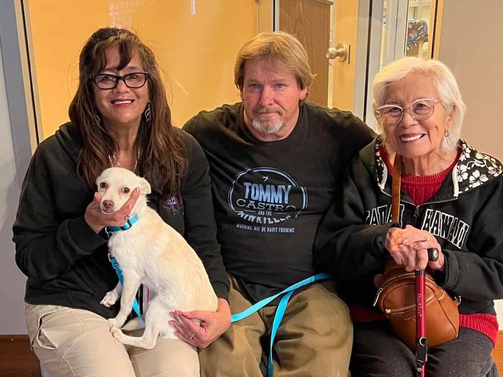 Three individuals seated together, smiling. A woman with glasses holds a small white dog. Next to her, a man wears a black shirt, and an older woman with silver hair sits on the right. The background is a bright indoor setting.