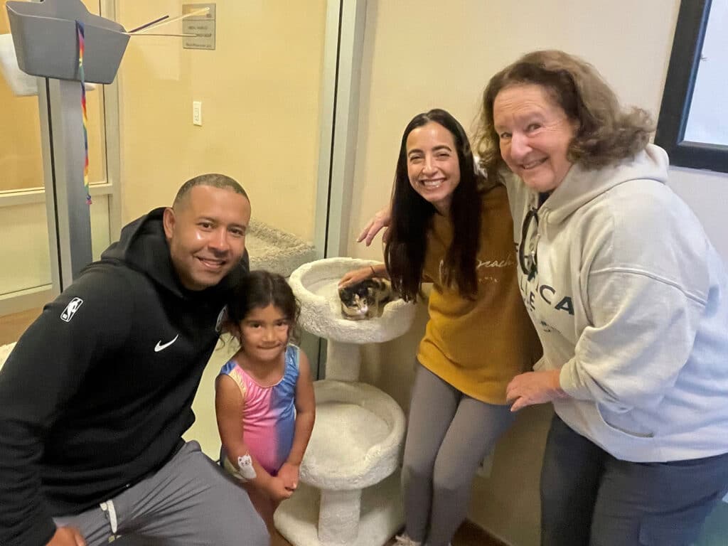 A man and a woman, along with a young girl and an older woman, smile while gathered around a cat in a shelter. The group stands near a cat tree, with the cat in the woman's lap. The setting appears to be an animal adoption center.