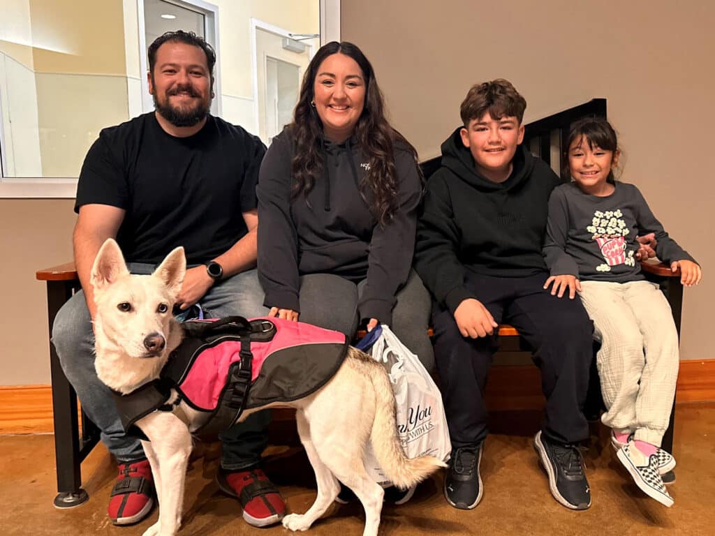 A family seated on a wooden bench, smiling at the camera. The group includes two adults and two children. A white dog with a pink vest sits beside them. The setting appears indoors, featuring neutral colored walls.
