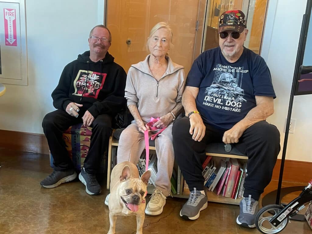 Three individuals seated indoors, with a light-colored French Bulldog sitting in front of them. The woman in the middle holds a leash attached to the dog. The first man wears a black hoodie, and the second man is in a blue t-shirt and sunglasses.
