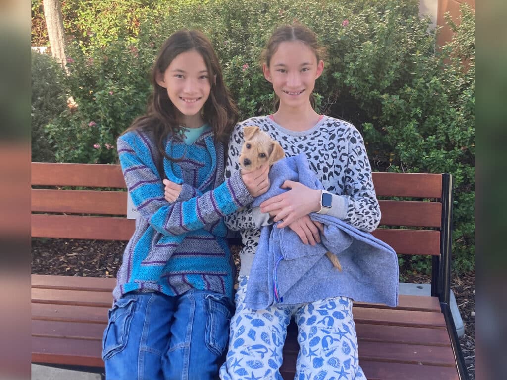 Two girls sit on a wooden bench outdoors, one wearing a blue and gray sweater and the other in a white sweater with a leopard print. The girl on the left holds a small, light-colored puppy wrapped in a gray blanket.