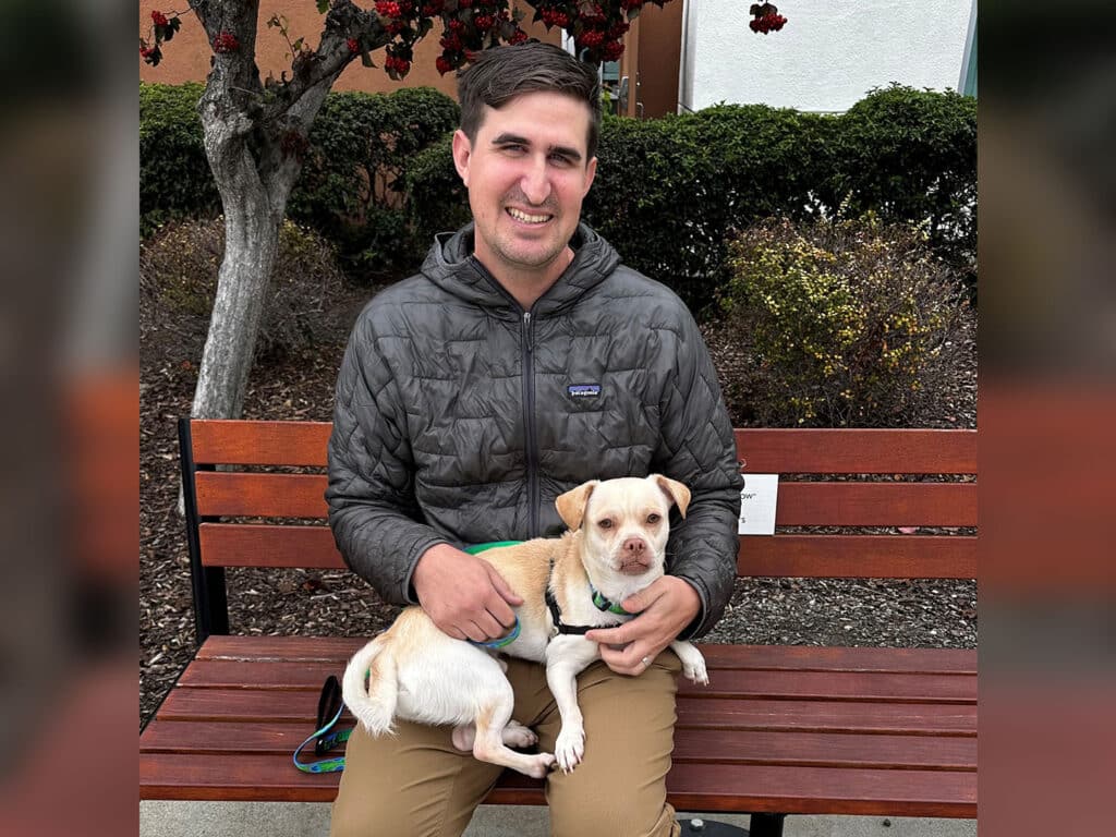 A man sitting on a wooden bench holds a small dog on his lap. He wears a gray jacket and light pants. The background includes a tree with red leaves and low green shrubs, indicating an outdoor setting.