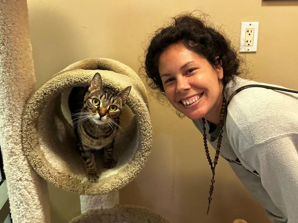 A woman smiling next to a tabby cat peeking out from a circular opening of a cat tree. The setting has a light-colored wall and a power outlet visible in the background.