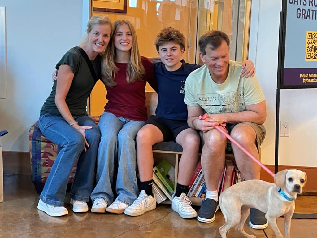 A family of four sits on a bench in an animal shelter. From left to right, a woman wearing a green shirt, a teenage girl in a maroon shirt, a teenage boy in a navy blue shirt, and a man in a light blue shirt. A small tan dog stands beside them on a leash.