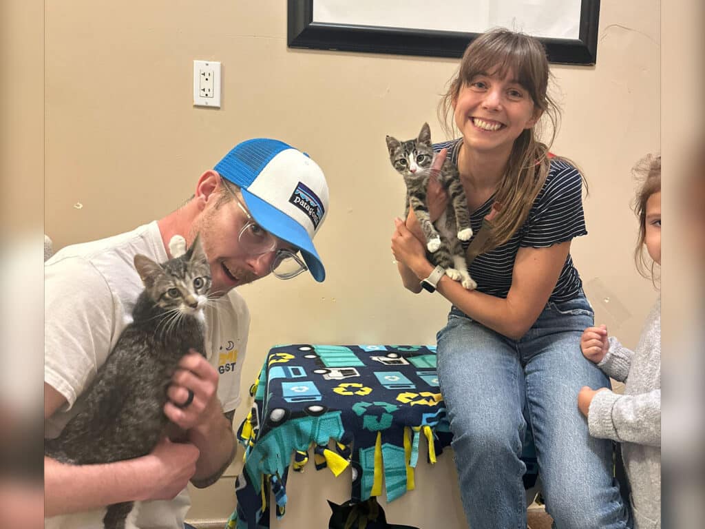A man wearing glasses and a blue cap holds a kitten, while a woman with long hair smiles and holds another kitten. A young girl is sitting beside them, looking at the camera. They are seated near a table covered with a fleece blanket in a warm setting.