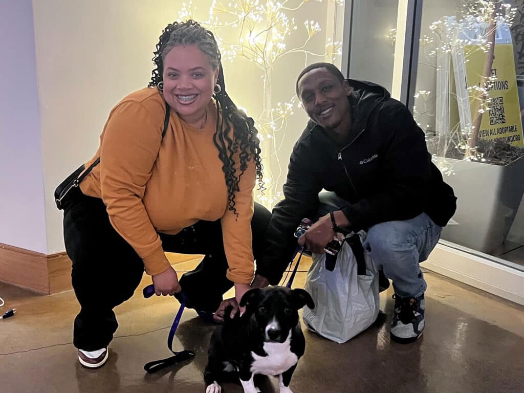 A woman with braided hair in an orange sweater and a man in a black jacket kneel together with a small black-and-white dog. They are inside a well-lit area with decorative lights in the background, smiling at the camera.
