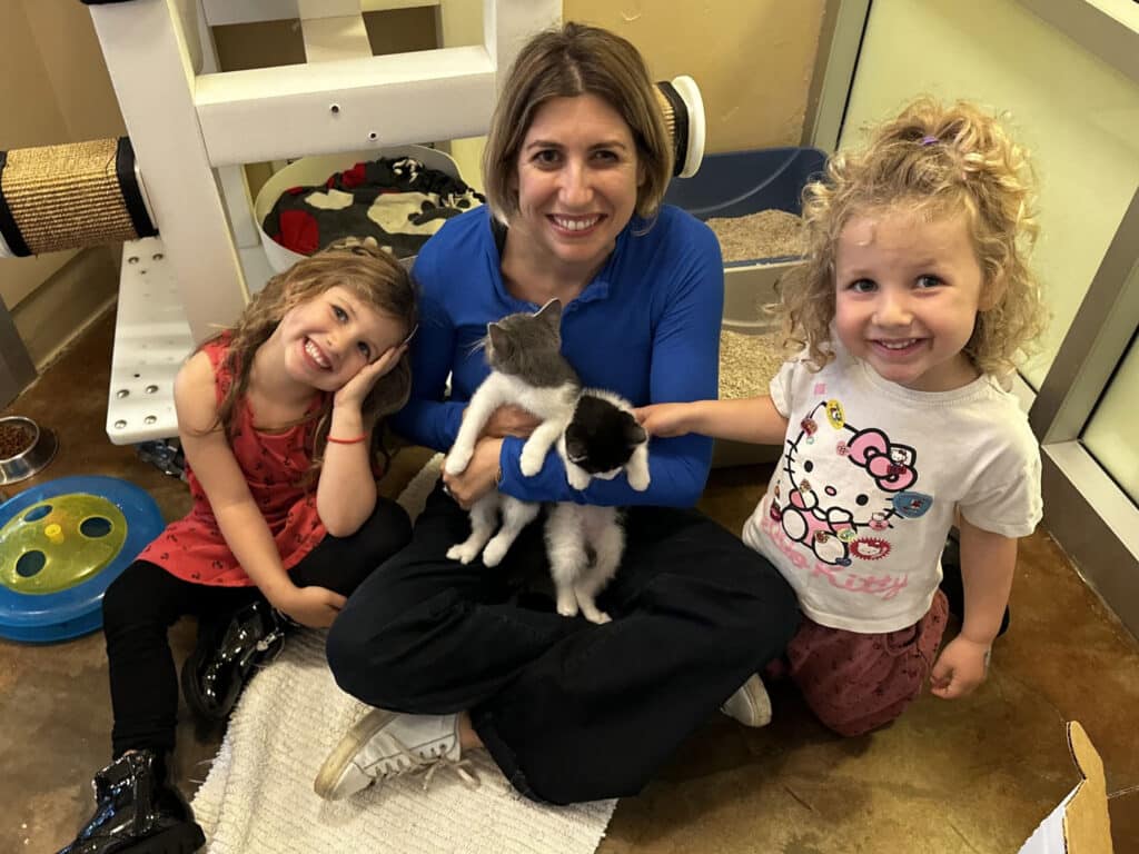 A woman wearing a blue shirt sits on the floor in a pet adoption center, holding two kittens. Two young girls, one in a striped shirt and the other in a Hello Kitty shirt, sit beside her, smiling. A cat tree and pet toys are visible in the background.