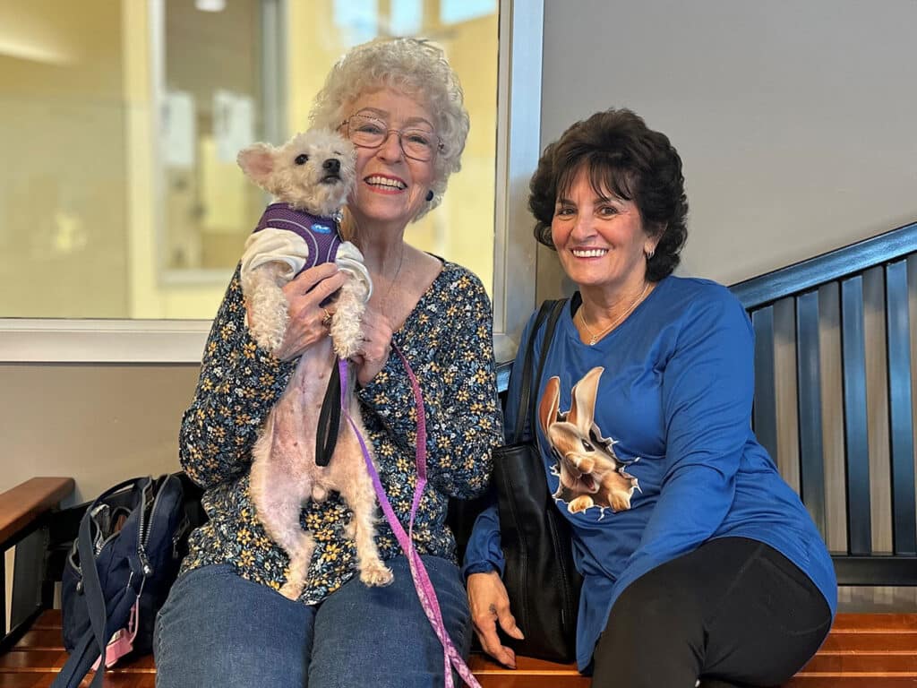 Two women sitting on a wooden bench inside a facility. The woman on the left holds a small dog and smiles, while the woman on the right poses alongside her. Both are casually dressed, with the background showing a light-colored wall and some indoor elements.