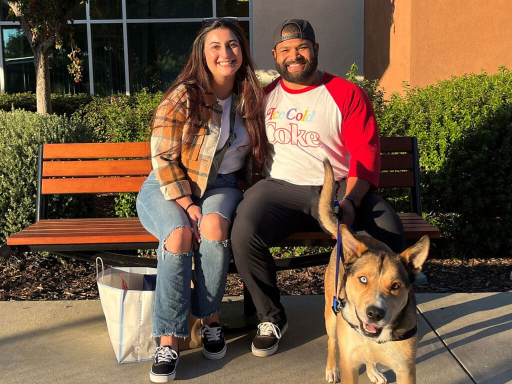 A woman and a man are sitting on a bench outside, smiling next to a medium-sized brown dog on a leash. The woman wears a plaid jacket and the man is in a shirt with a logo. A shopping bag is on the ground beside them.