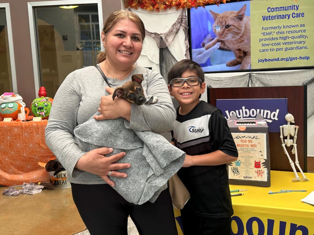 A woman holds a small animal wrapped in a blanket while a young boy smiles beside her. They are in an indoor space decorated for an event, with a banner in the background promoting community veterinary care and adoption services.