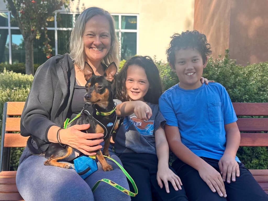A woman smiles while sitting on a bench with a small dog on her lap. Next to her, a girl and a boy sit closely, pointing and smiling. They are outdoors in a green setting, with trees and a building visible in the background.