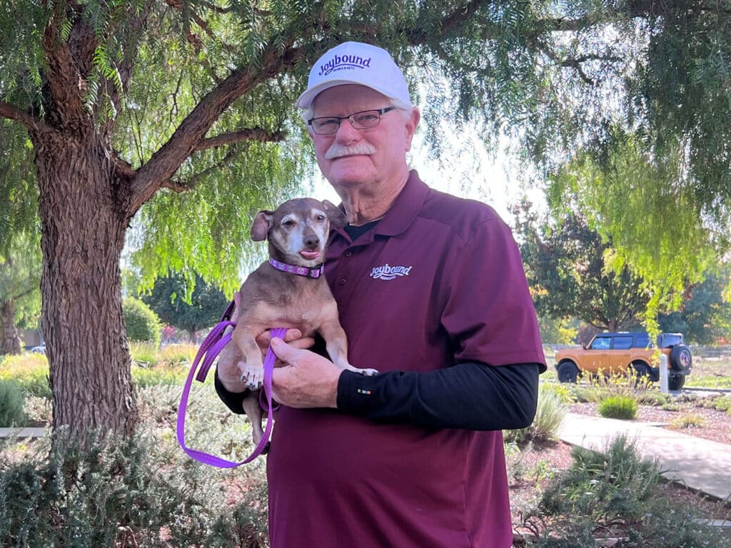 An older man wearing a baseball cap and a maroon shirt holds a small dog in his arms. The background features greenery, a tree, and a vehicle partially visible behind them, suggesting an outdoor setting.