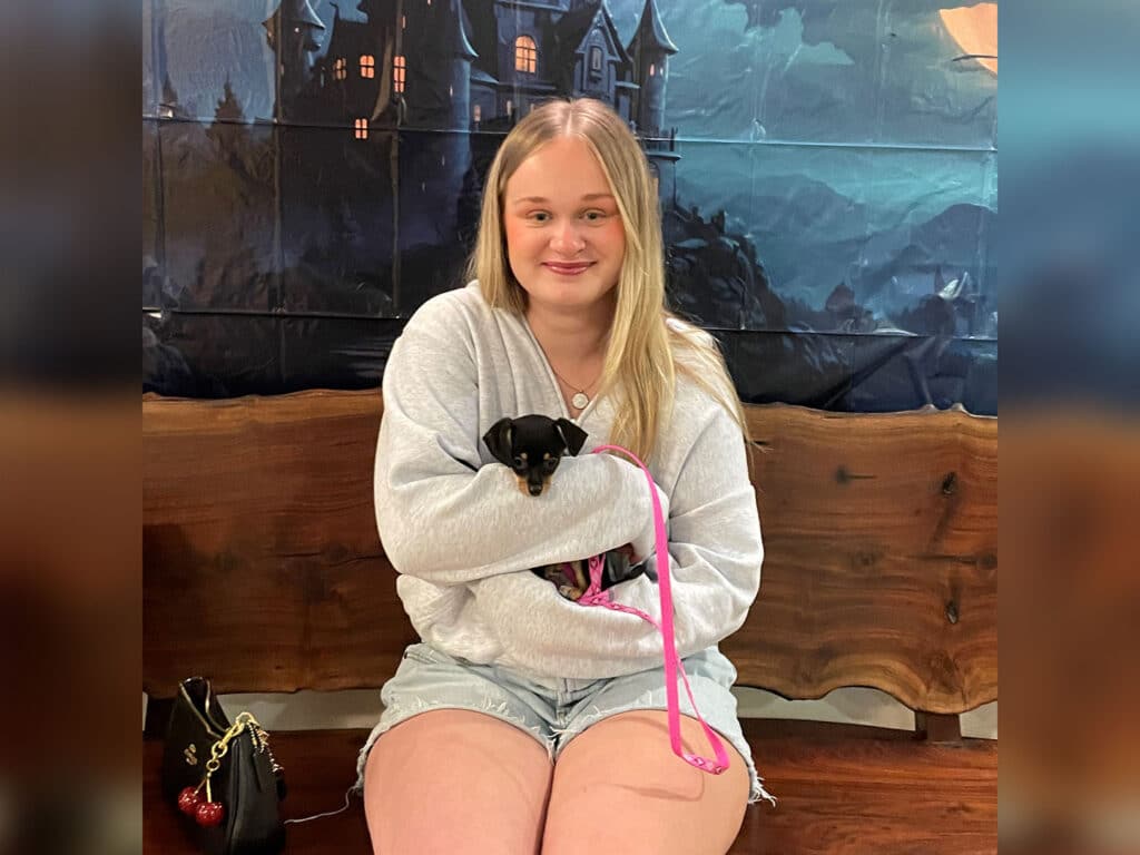 A young woman sits on a wooden bench with a small black dog in her arms, wearing a light gray sweater and denim shorts. The background features a mural with a castle and mountains, creating a whimsical atmosphere.