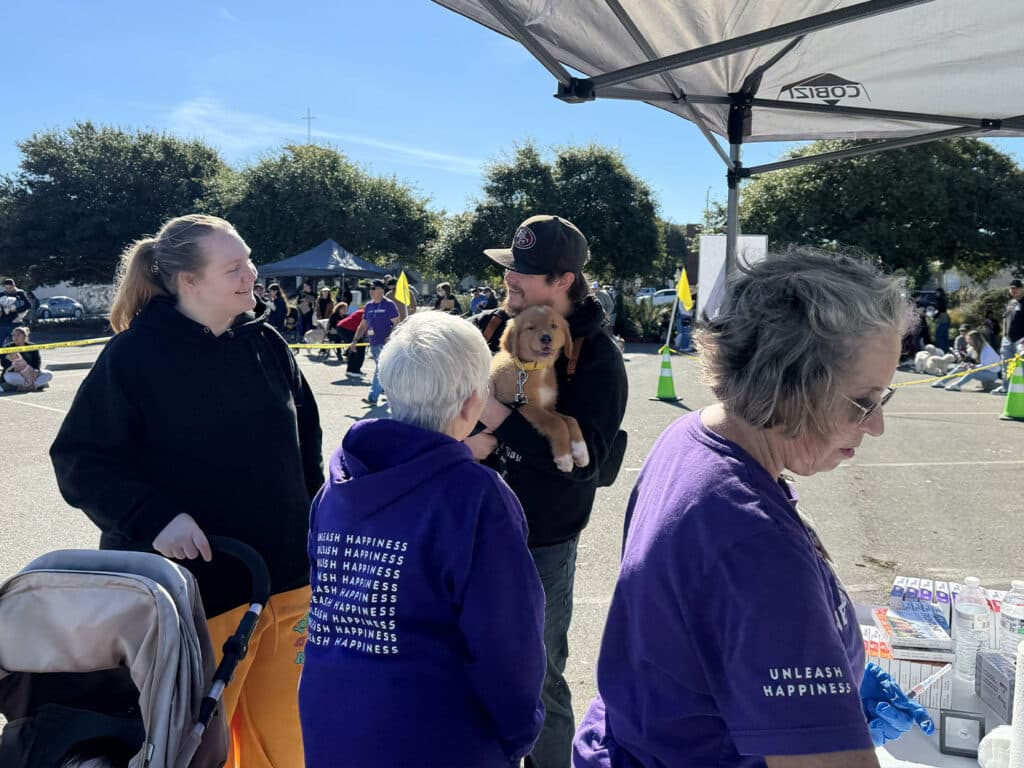 At a community event, a man holds a small dog while talking to a woman in a black hoodie. Two other women, one in a purple sweatshirt and another with glasses, are at a table with various supplies, with people gathered in the background.