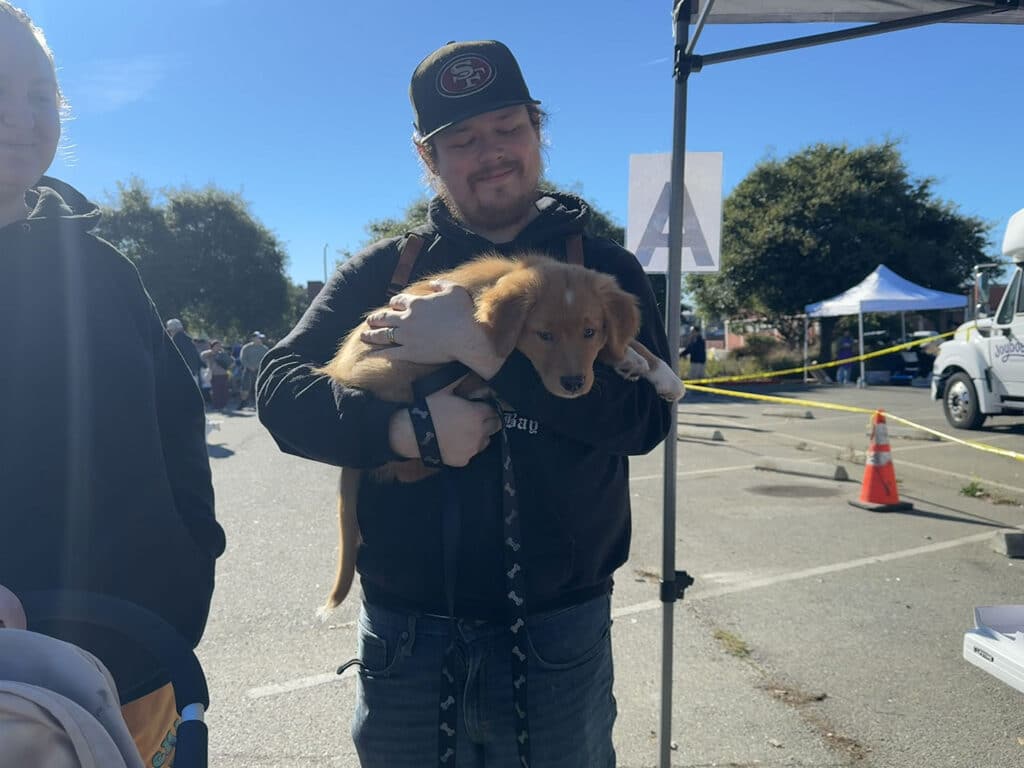 A person holding a golden retriever puppy in their arms, smiling while standing at an outdoor event. The background shows a parking lot with people, trees, and tents. Colorful cones are visible on the ground.