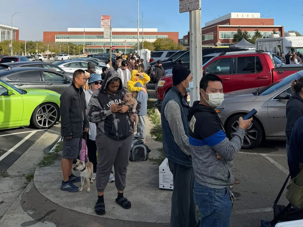 A line of people waiting outside a vaccination clinic in a parking lot, with a mix of individuals wearing casual clothing. A small dog is present, and cars are parked in the background. The setting appears to be during daylight.
