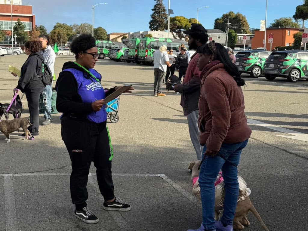 A community member wearing a blue vest stands with a clipboard, speaking to two individuals in a parking lot. One person holds a small dog, and another individual is in a brown jacket. In the background, vehicles are visible.