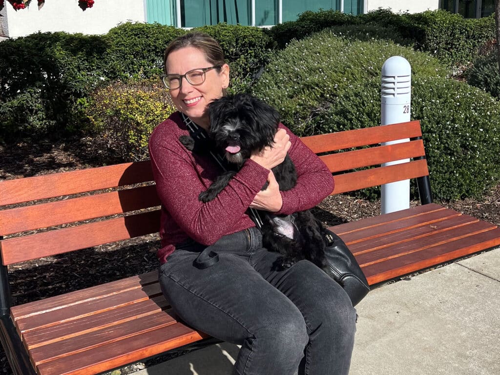 A woman with glasses sits on a bench outdoors, holding a small black dog. The scene features greenery in the background, and sunlight casts soft shadows, creating a warm atmosphere.