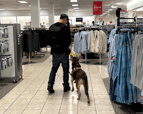 A man walking in a department store with a service dog. The dog is wearing a harness and walking beside him on a leash. Clothing racks are visible on both sides, with a bright, spacious store interior in the background.