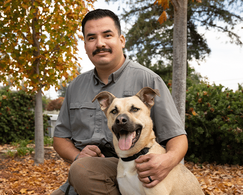 A man with a mustache sits on the ground next to a light-colored dog, both looking towards the camera. The background features autumn foliage and greenery, creating a warm outdoor setting.