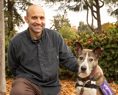 A man with short hair smiles while sitting beside a service dog wearing a purple harness. They are in a park setting with trees and fall foliage in the background.