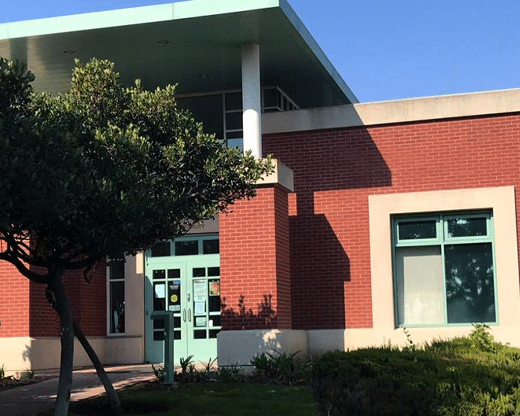 Exterior view of a brick building with large windows and a light green roof. A tree stands near the entrance, which has glass doors and is framed by small shrubs and grass. The scene is set under a clear blue sky.