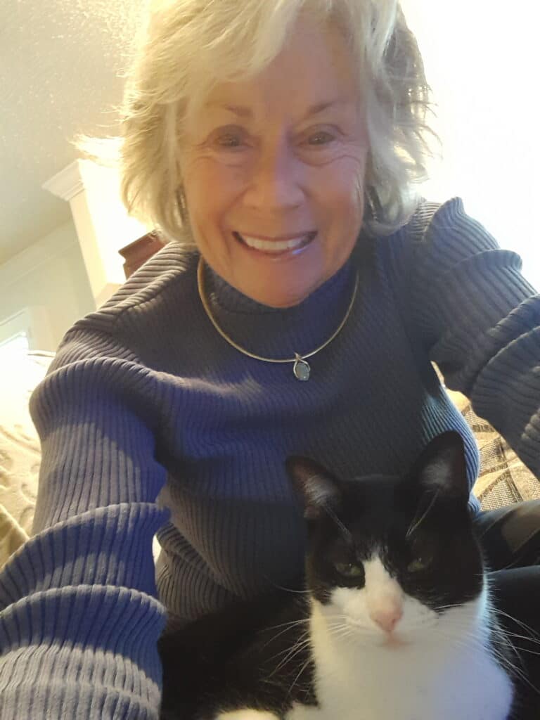 A woman with gray hair wearing a blue sweater smiles while taking a selfie. A black and white cat sits beside her, partially in her lap. The setting appears to be indoors, with soft lighting and a cozy atmosphere. Relevant to the Joybound Strategic Plan.