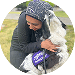 A person wearing a headscarf smiles and embraces a dog, both sitting on grass. The dog wears a purple vest indicating its role, illustrating the bond between the volunteer and the animal.