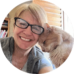 A smiling woman wearing glasses sits next to a light-colored cat with its head resting against her. The background includes shelves with books, suggesting a cozy indoor environment.