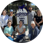 A group of seven volunteers posing in front of a sign for 'Solo's Play Ground' in a park. They stand in a semi-circle, smiling. The sign features a blue and white design with an image of a dog. The setting is sunny and green.