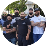 A group of six men with beards, wearing black and white t-shirts, pose together outdoors in front of a fountain. They are standing with their arms crossed and displaying a confident stance, emphasizing camaraderie among volunteers.