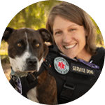A woman smiles while posing with a brown and white dog wearing a service vest. The background features greenery, suggesting an outdoor setting. Both the volunteer and the dog appear relaxed and friendly.