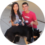 A young woman and a young man smiling while holding the leash of a black dog. They are both wearing name tags and are standing on a sidewalk in a sunny outdoor setting, representing volunteers engaging with animals.