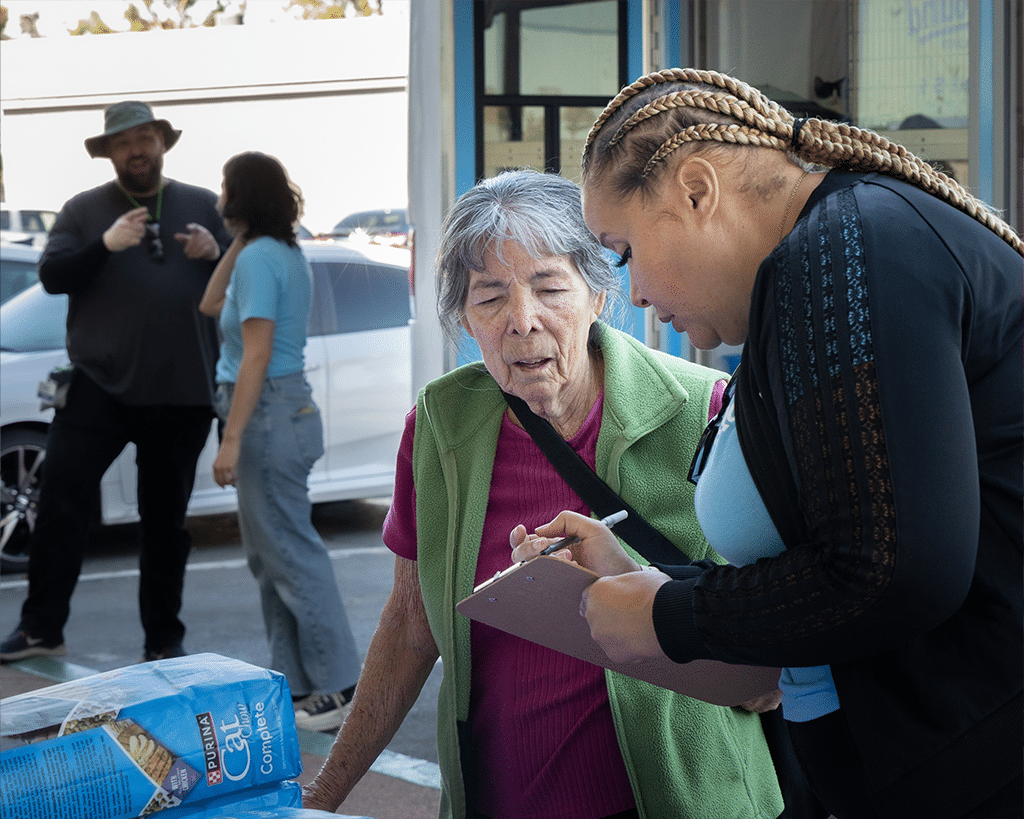 A woman with braided hair assists an elderly woman in a green vest while taking notes on a clipboard. In the background, a man stands near a car, looking towards them. The setting appears to be an outdoor admissions event.