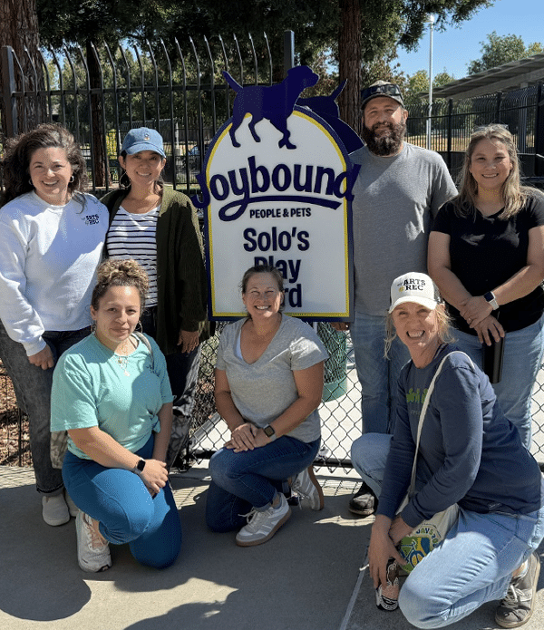 A group of eight volunteers posing in front of a sign that reads "Joybound People & Pets Solo's Play Yard". They are smiling and dressed casually, with a mix of hairstyles and attire, surrounded by a fence and trees.