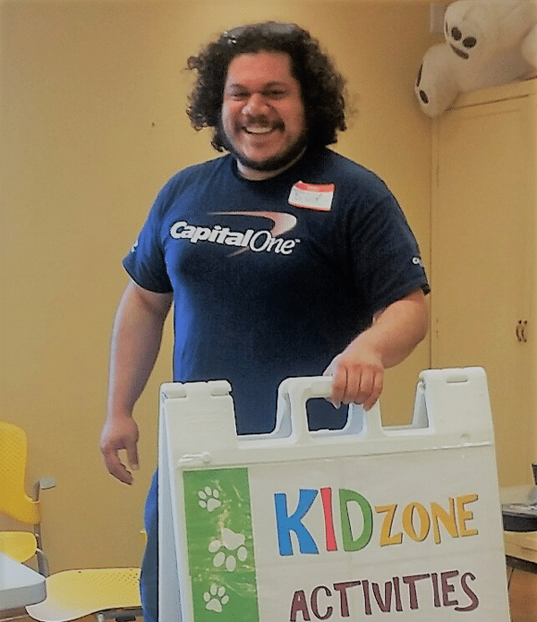 A smiling volunteer wearing a Capital One t-shirt stands next to a sign that reads 'KIDZONE ACTIVITIES' in a classroom setting with yellow walls and chairs.