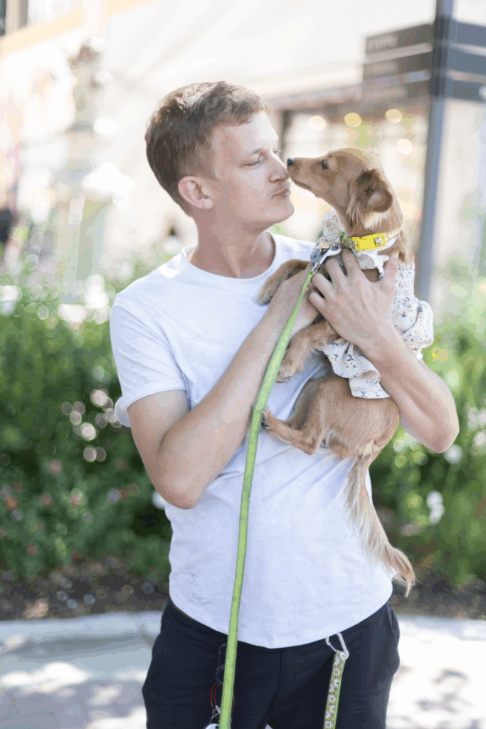 A man wearing a white t-shirt holds a small dog in his arms, with the dog leaning towards his face. They are surrounded by greenery and soft blurred lights in the background, contributing to a relaxed outdoor atmosphere. This image relates to the Joybound Strategic Plan.