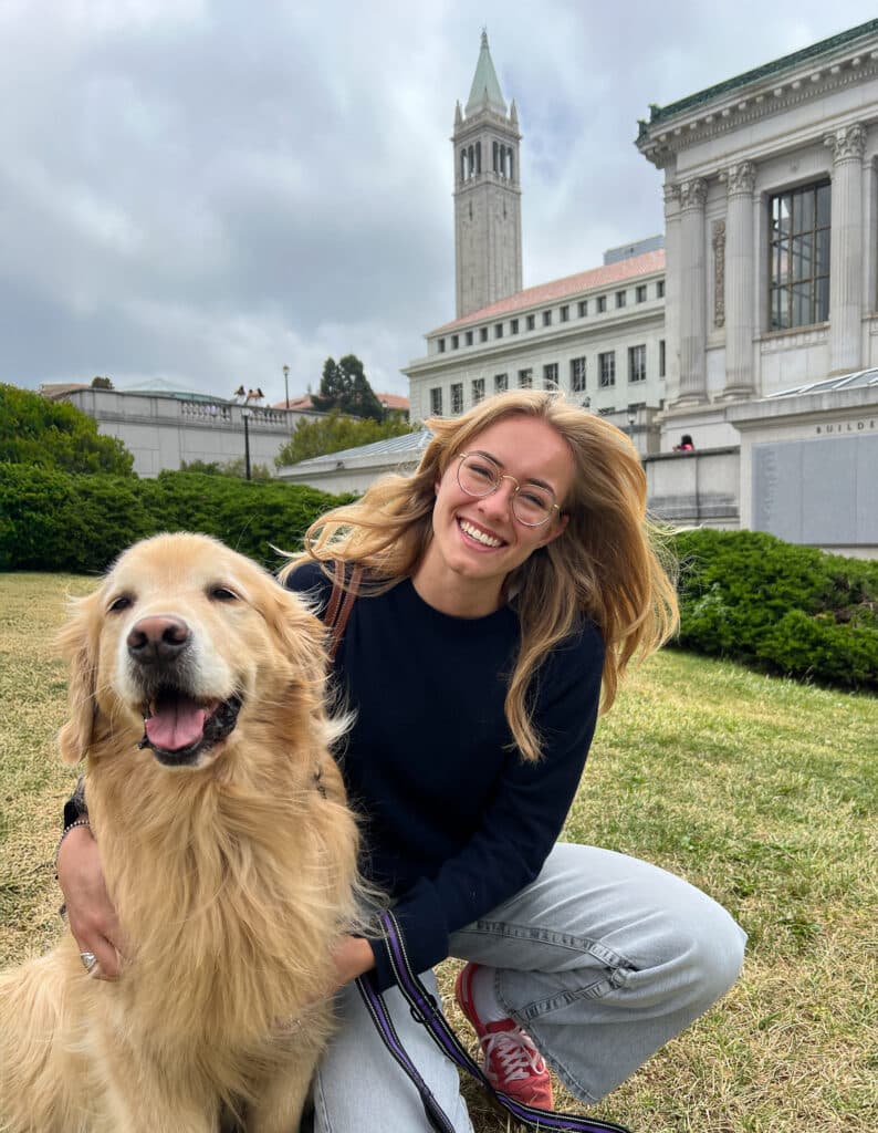 A woman with glasses sits on grass next to a golden retriever, smiling at the camera. In the background, a tall clock tower and historic building are visible. This image supports the Joybound Strategic Plan within the Impact Report 2024-2025.
