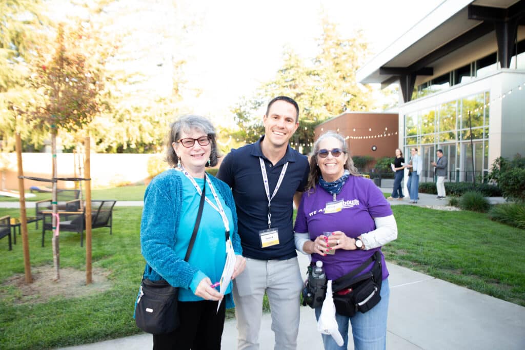 Three individuals pose together outdoors during an event. Two women, one wearing glasses and a blue sweater, and another in a purple shirt, stand on either side of a man in a black shirt. The setting includes greenery and a modern building.