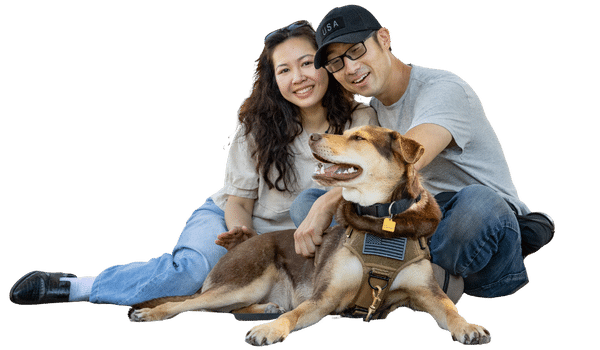 A woman and a man are sitting on the ground with a dog in a harness. The dog is looking up and smiling, while the couple smiles at the camera. The image supports the Joybound Strategic Plan outlined in the Impact Report 2024-2025.