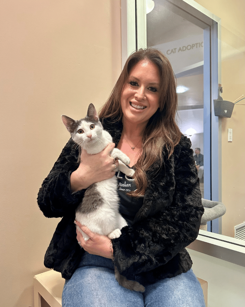 A woman in a black fur coat smiles while holding a gray and white cat in her arms. They are in a cat adoption center, with a beige wall background and a light-colored area visible.