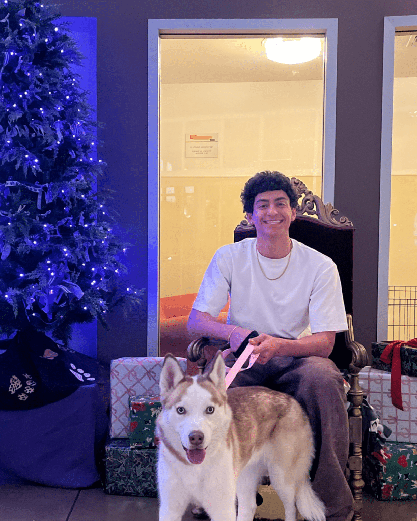 A young man sitting on a throne-like chair with a friendly Siberian Husky by his side. The man is wearing a white shirt and has a slight smile. Behind them is a decorated Christmas tree and wrapped gifts.