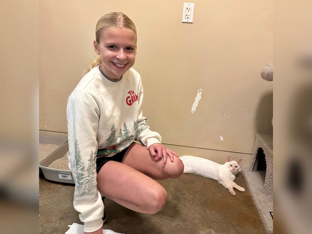 A girl with long blonde hair, wearing a festive sweatshirt, smiles while kneeling beside a lounging white cat in a room with a litter box. The cat appears relaxed on the floor near a small entrance.