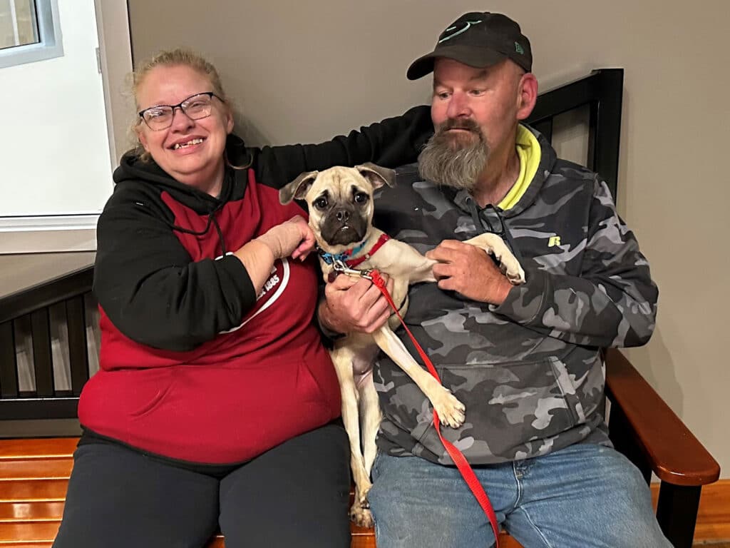 A couple sitting on a wooden bench inside a building, smiling while holding a small dog. The woman wears glasses and a red sweatshirt, while the man, with a beard, is in a camouflage jacket. The dog is on a leash, appearing relaxed between them.
