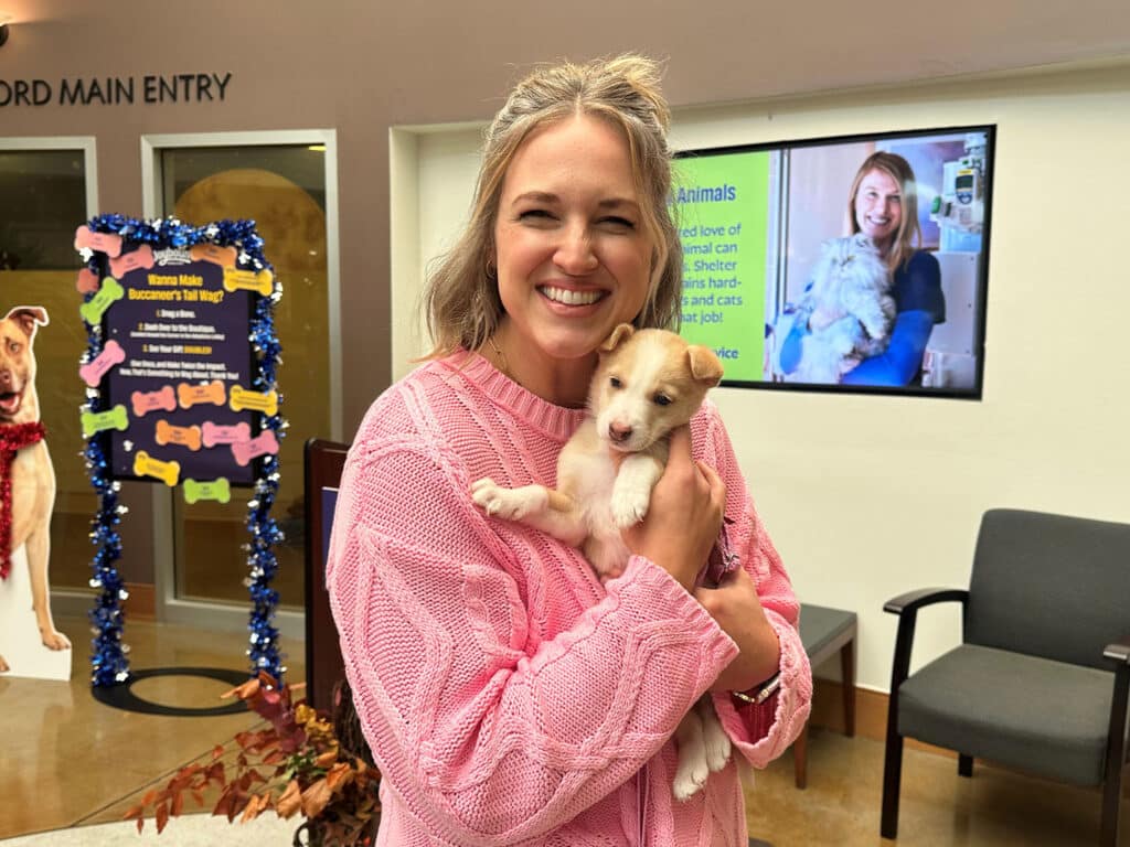A woman wearing a pink sweater smiles while holding a small puppy. In the background, a digital screen displays an image of another woman with a dog. The setting appears to be an animal shelter or adoption center.