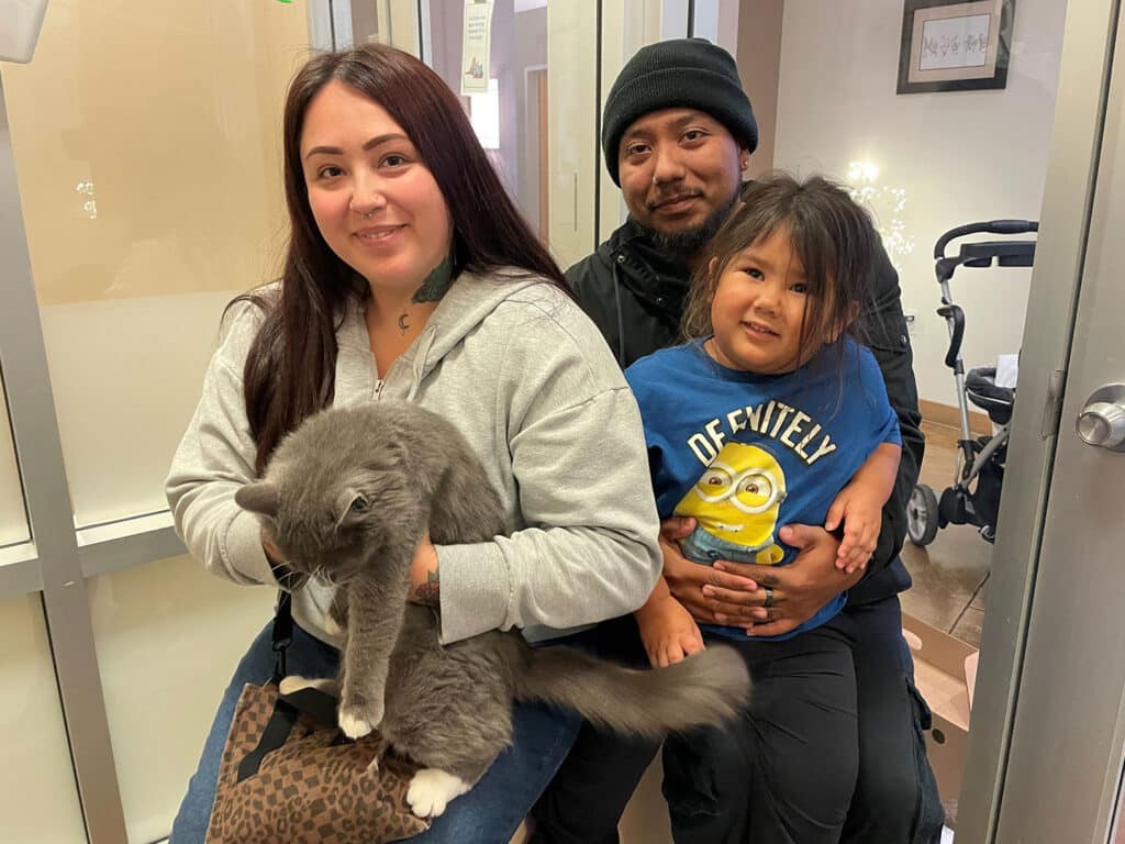A woman holds a gray cat while sitting next to a man who has a young girl on his lap. The family is in an indoor setting with a stroller visible in the background. The mood appears relaxed.